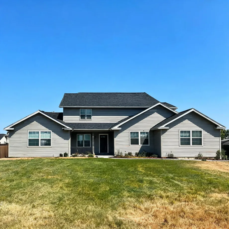 A two-story house with light gray horizontal siding, dark gray shingled gable roofs and a dark front door, fronted by a large partly browned lawn under a clear blue sky.