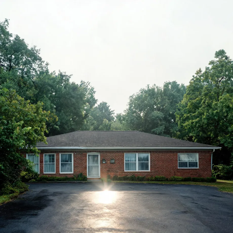 A single-story red brick building with a dark hipped roof, white-framed windows and a white front door, fronted by a large asphalt parking area with sun glare, surrounded by dense green trees under a pale sky.