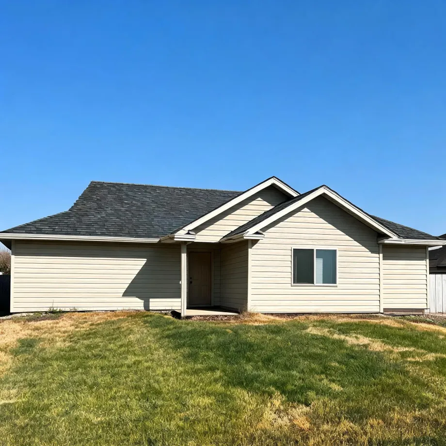 Single-story house with beige horizontal lap siding and a dark gray shingled gable roof, with a single window and a recessed front door, fronted by a patchy green and brown lawn under a clear blue sky.