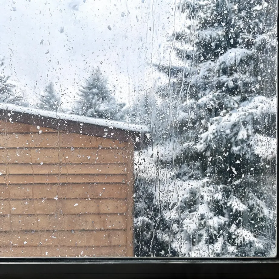 View through a window covered in water droplets and streaks, showing a wood-sided building with a snow-topped roof and snow-laden conifer trees under a pale sky.