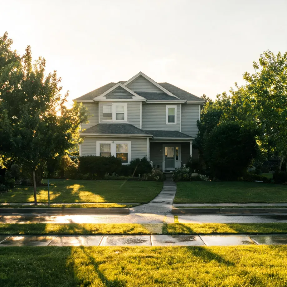A two-story house with light gray horizontal siding and a covered front porch, seen from across a wet street with a lawn, sidewalk, shrubs and leafy trees in low sunlight.