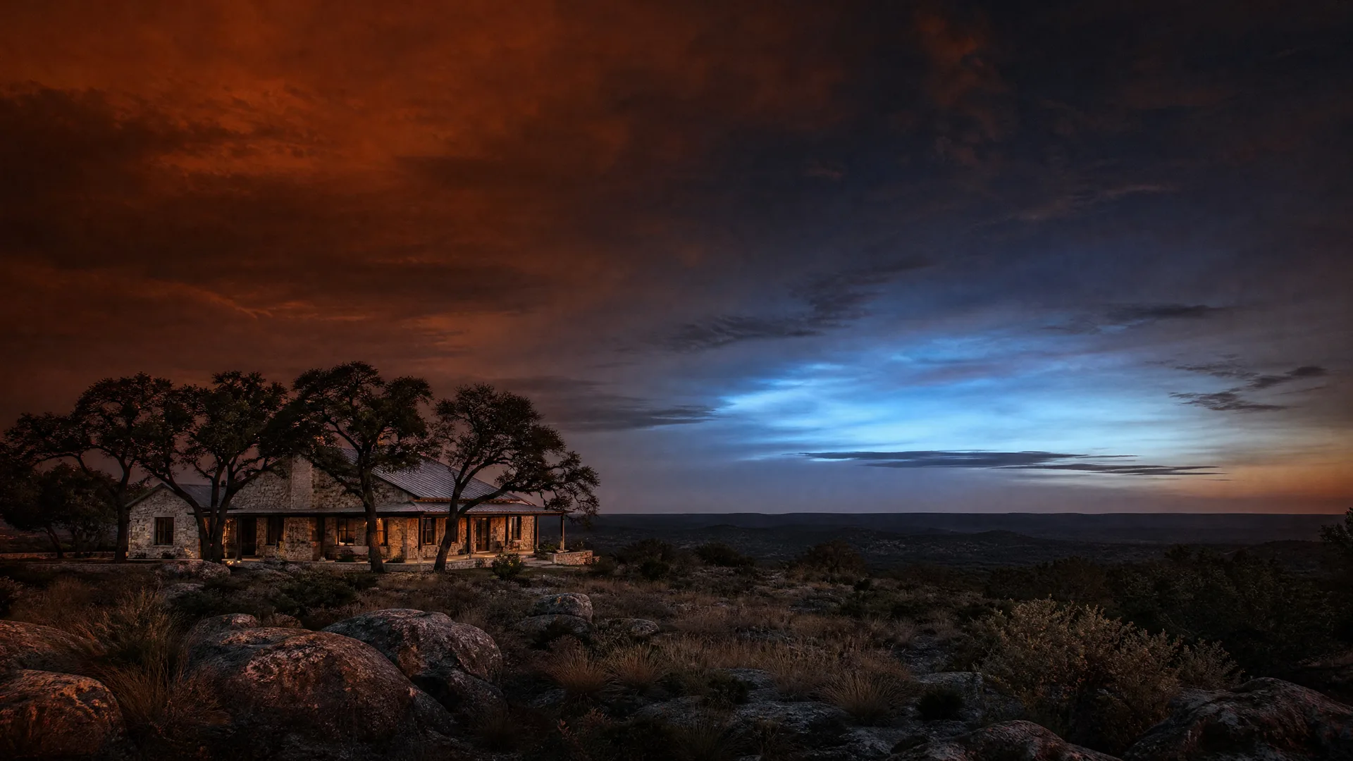 A stone house with a metal roof and covered porch sits among oak trees on a rocky hilltop at dusk, with scrub brush in the foreground and a glowing blue and orange cloudy sky above a distant flat horizon.