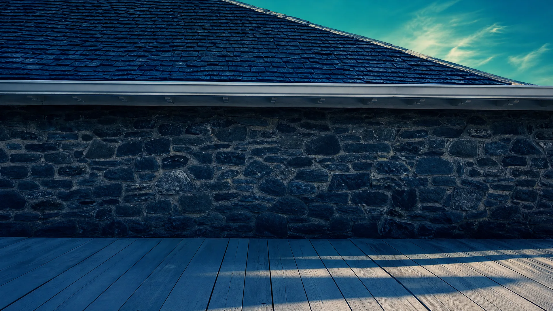 A dark stone wall with a shingled roof and white eave runs across the frame above a gray wood plank deck with diagonal shadows, under a blue sky.