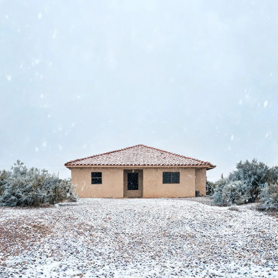 A single-story tan stucco house with a red tile hip roof stands behind snow-dusted gravel ground and frosted shrubs during falling snow under a pale sky.