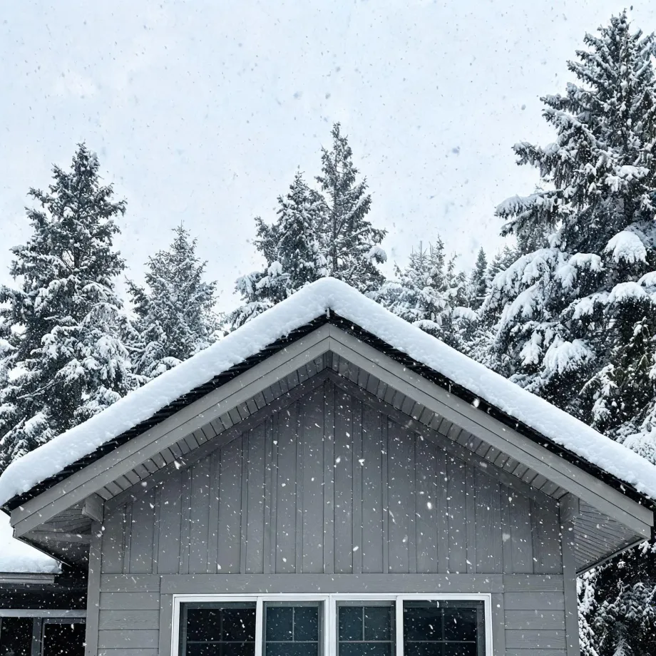 The snow-covered gable end of a gray board-and-batten house with wide eaves and a window below, backed by snow-laden conifers under a pale sky with falling snowflakes.