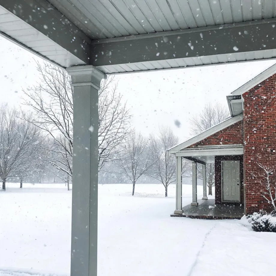 View from under a covered porch with a white column toward a red brick house wing with a gray door and small porch, surrounded by snow-covered ground and bare trees during falling snow.