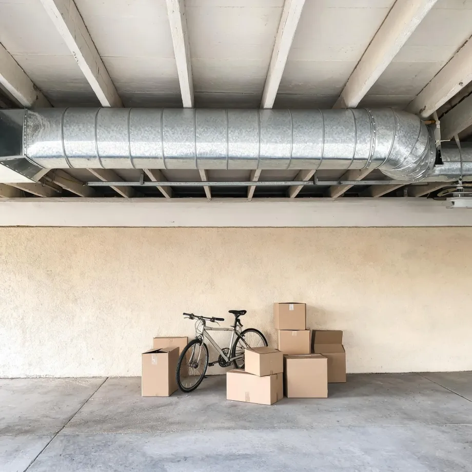 A bicycle leaning against a beige stucco wall surrounded by several cardboard boxes in a covered parking area, with a large metal duct running beneath the concrete beam ceiling.
