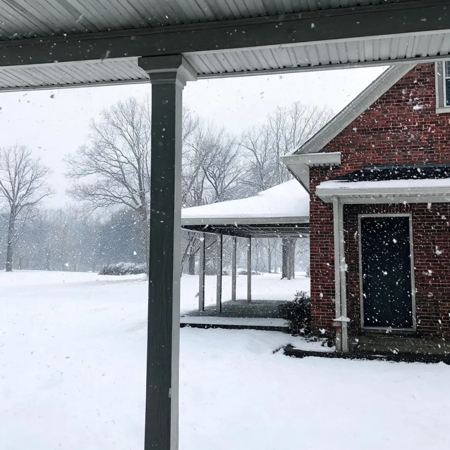View from under a covered porch during falling snow, showing a snow-covered lawn, bare trees, and a brick building with a dark door and a curved covered walkway.