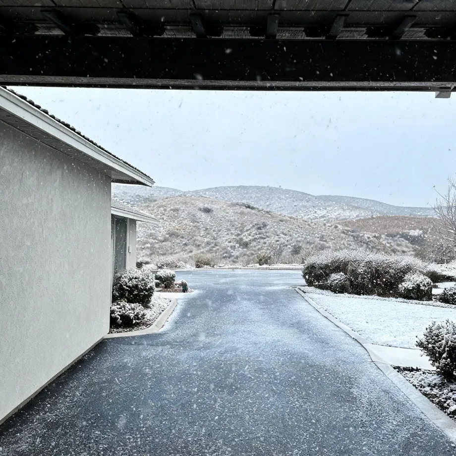 View from under a covered carport of a paved driveway beside a white stucco house with tile roof, with snow-dusted shrubs, lawn and hills in the background during falling snow.