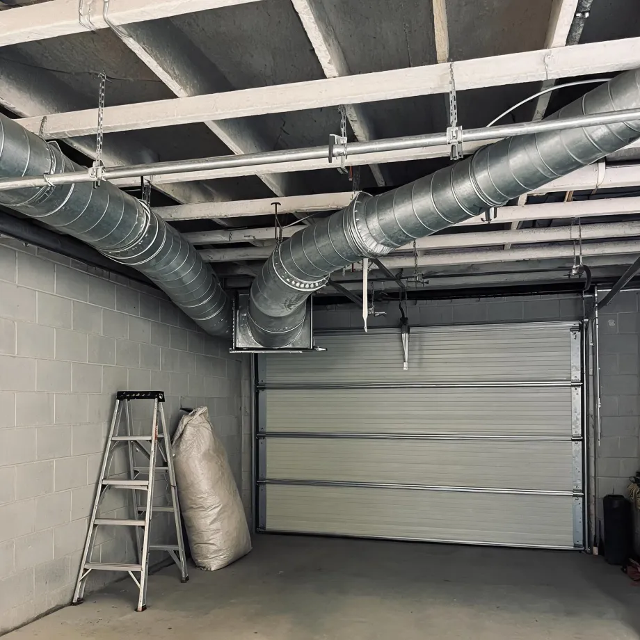 Interior of a concrete block garage with a closed sectional door, exposed ceiling joists and suspended round galvanized ductwork, a step ladder and a large white bag against the left wall.