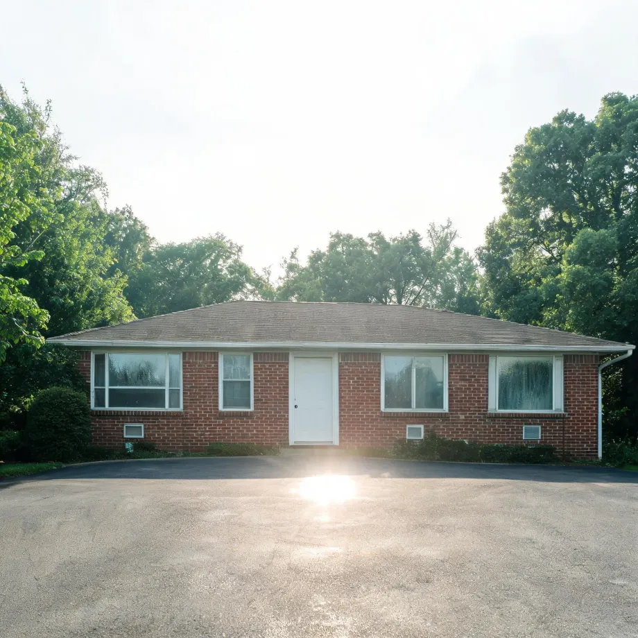 A single-story red brick ranch house with a white front door, large windows and a shingled roof, fronted by a wide paved lot with sun flare, surrounded by green deciduous trees.