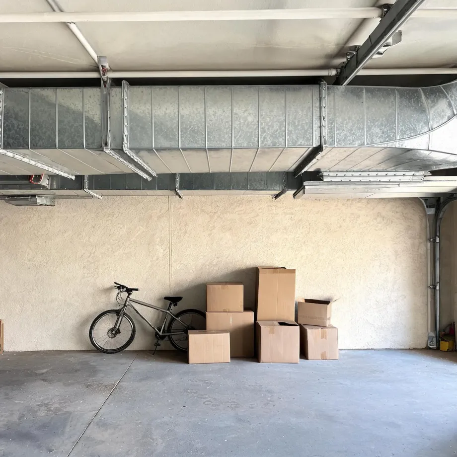 Interior of a garage with exposed galvanized ductwork below the ceiling, a stucco wall, a bicycle leaning against the wall, and several stacked cardboard boxes on a concrete floor.