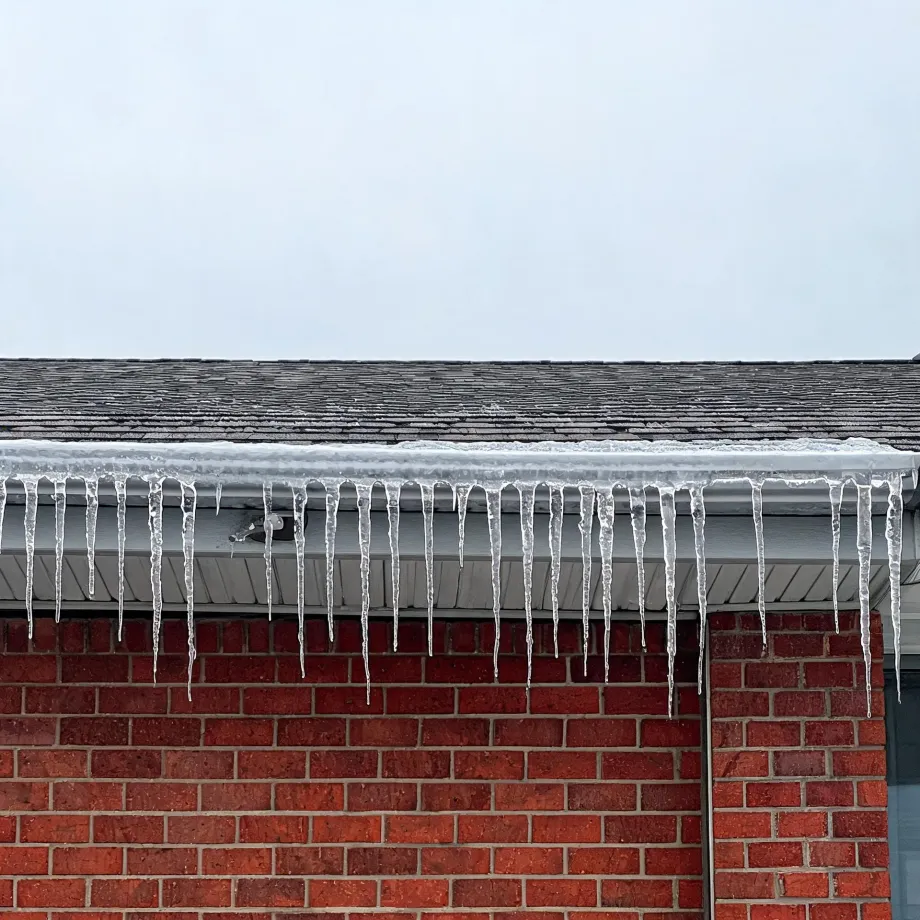 Icicles hang from a snow-covered gutter along the eave of a red brick house with a shingled roof under an overcast sky.