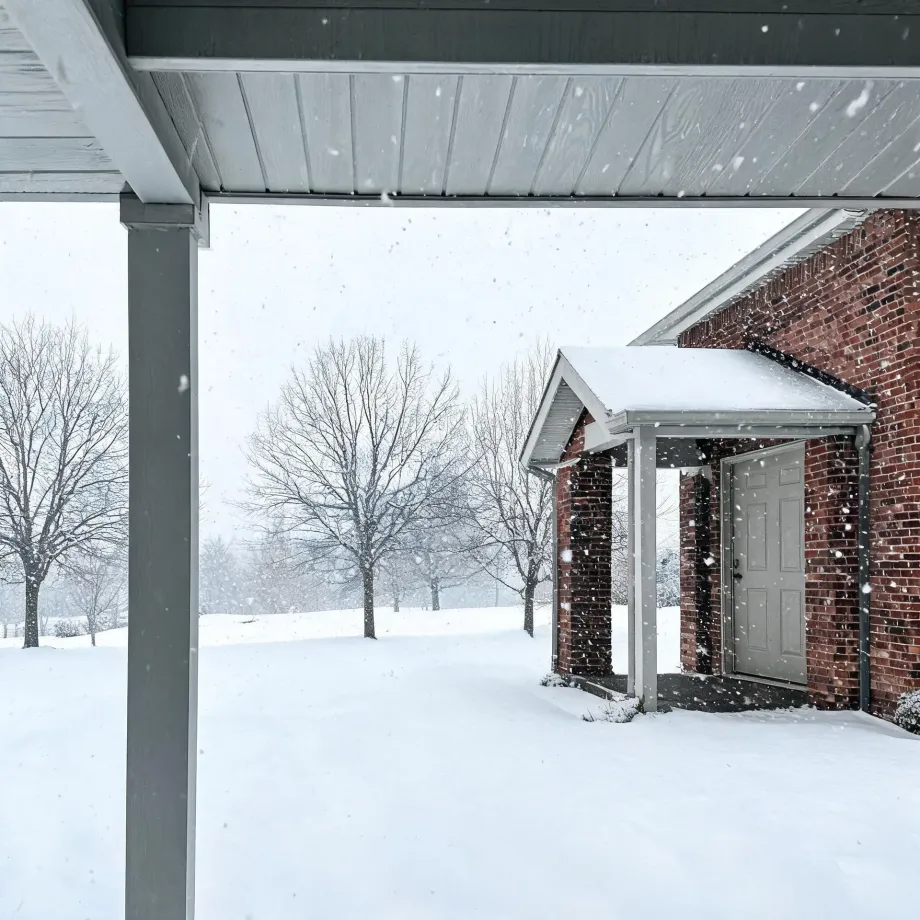 View from under a covered porch toward a red brick house with a gray side door beneath a small gabled entry canopy, with falling snow, snow-covered ground and bare trees in the background.