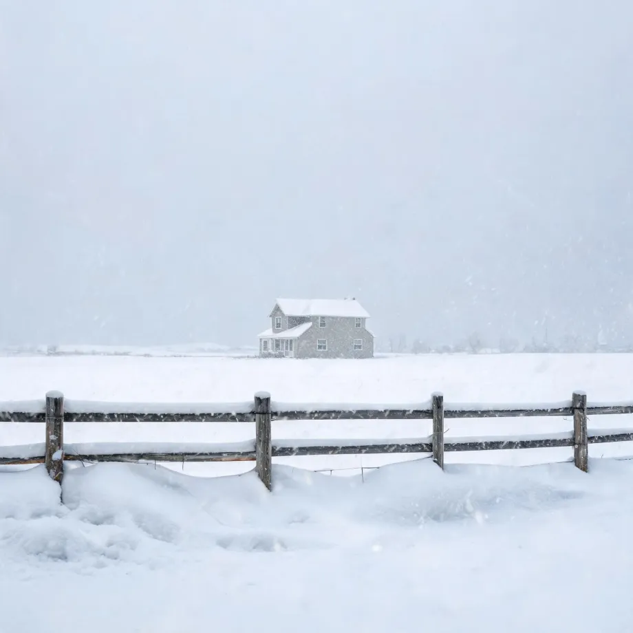 A two-story gray house with a snow-covered roof stands alone in a flat snow-covered field during falling snow, with a weathered wooden post-and-rail fence in the foreground.