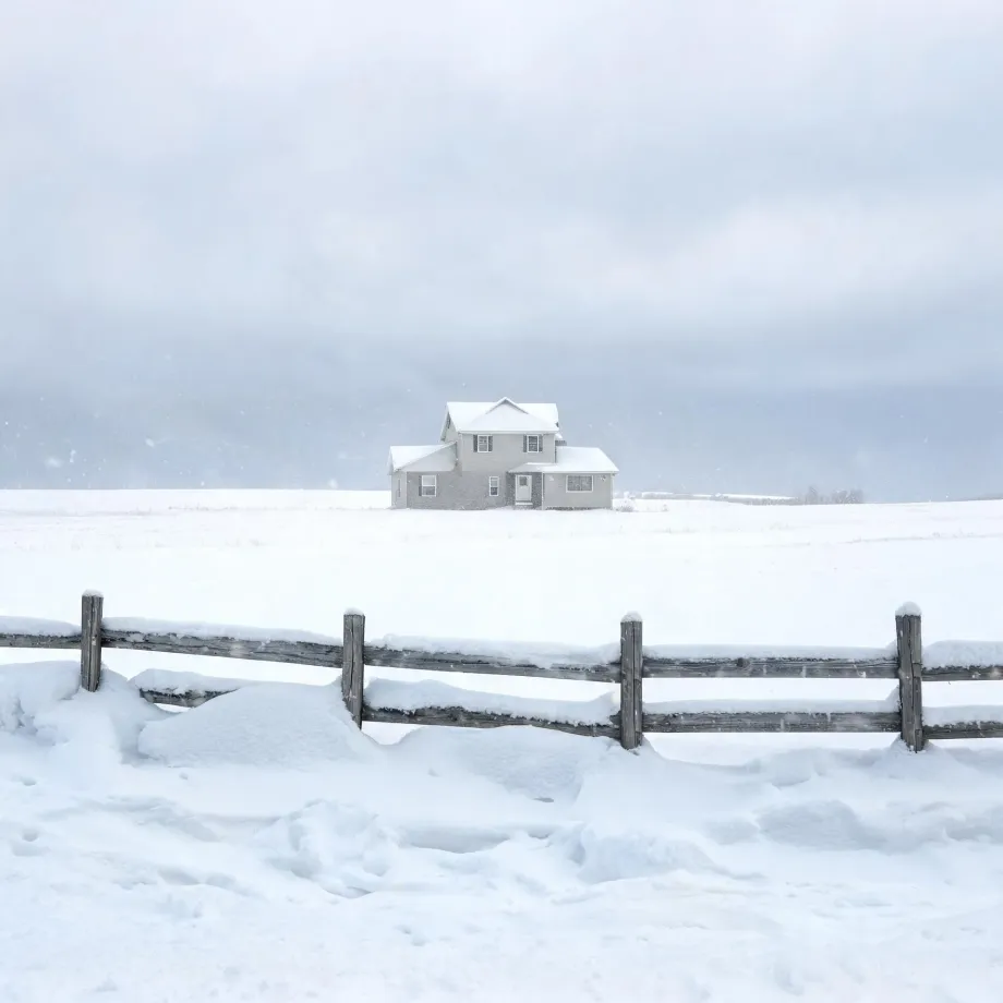 A two-story light-colored house stands alone in a snow-covered field under an overcast sky, with a snow-laden wooden rail fence crossing the foreground.