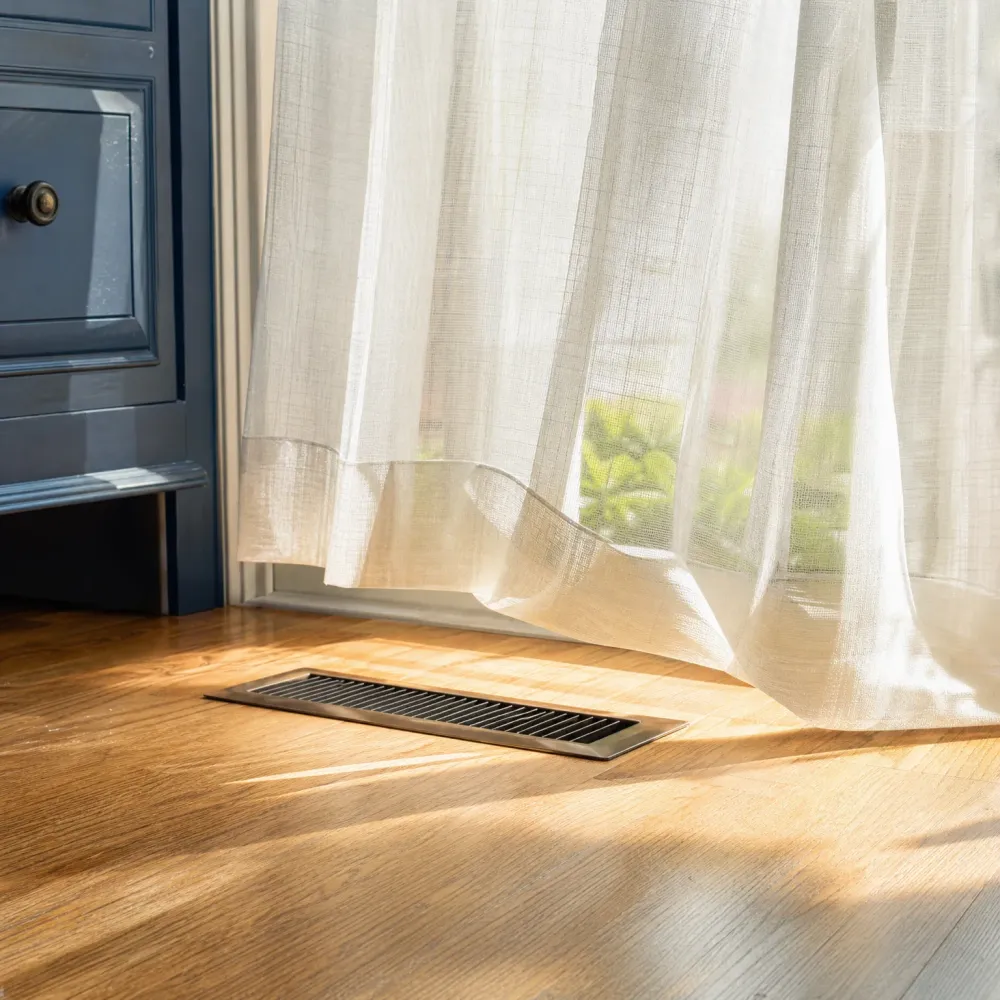 A metal floor vent set in oak flooring beside a sheer white curtain and a dark blue cabinet, with sunlight casting shadows across the floor.