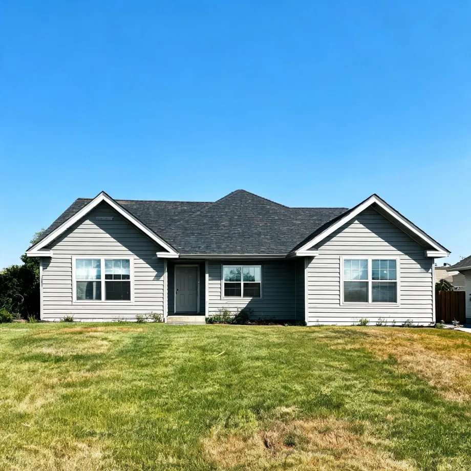 Single-story gray horizontal-siding house with two front gables, dark shingle roof, white-trimmed windows and a gray front door, fronted by a patchy green lawn under a clear blue sky.