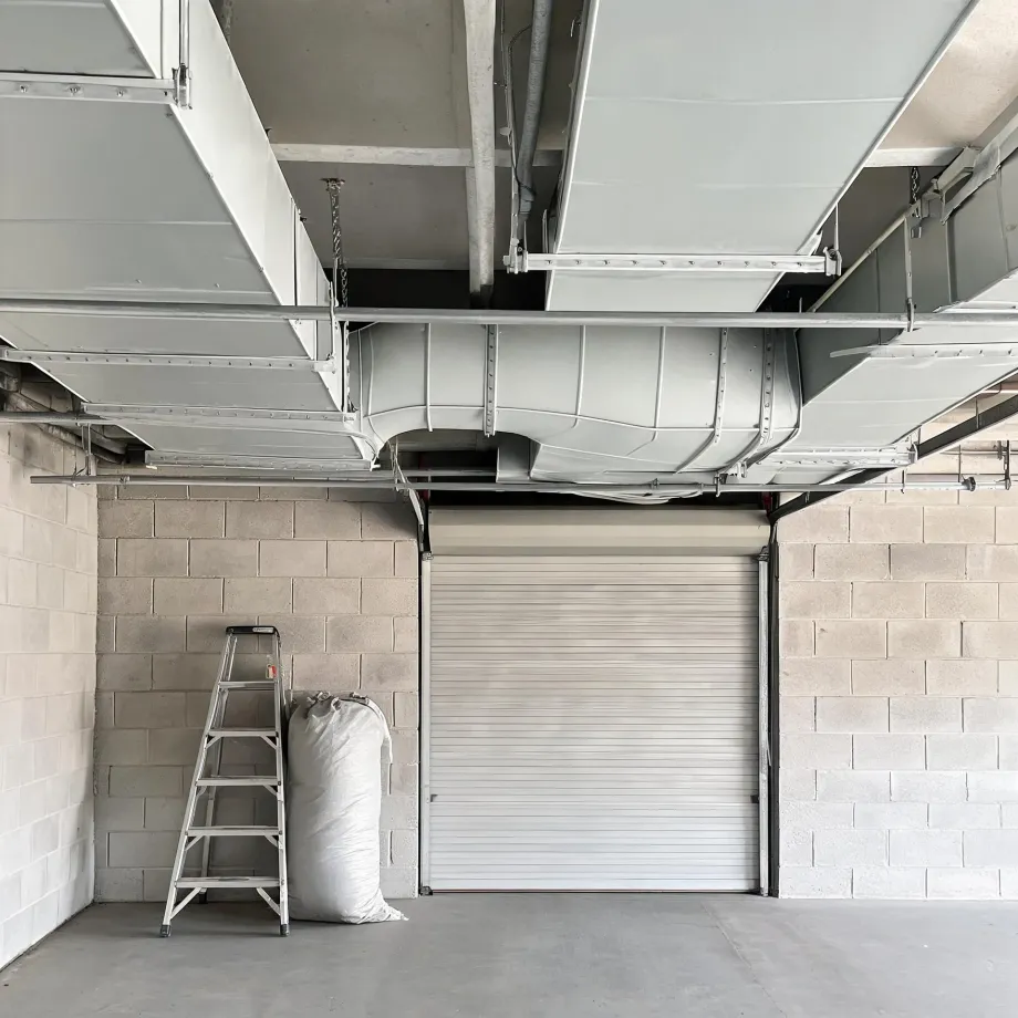 Interior of a concrete block garage with a closed white roller shutter door, a folding aluminum stepladder and a filled white sack against the left wall, and large light-gray sheet metal ducts running across the exposed ceiling.