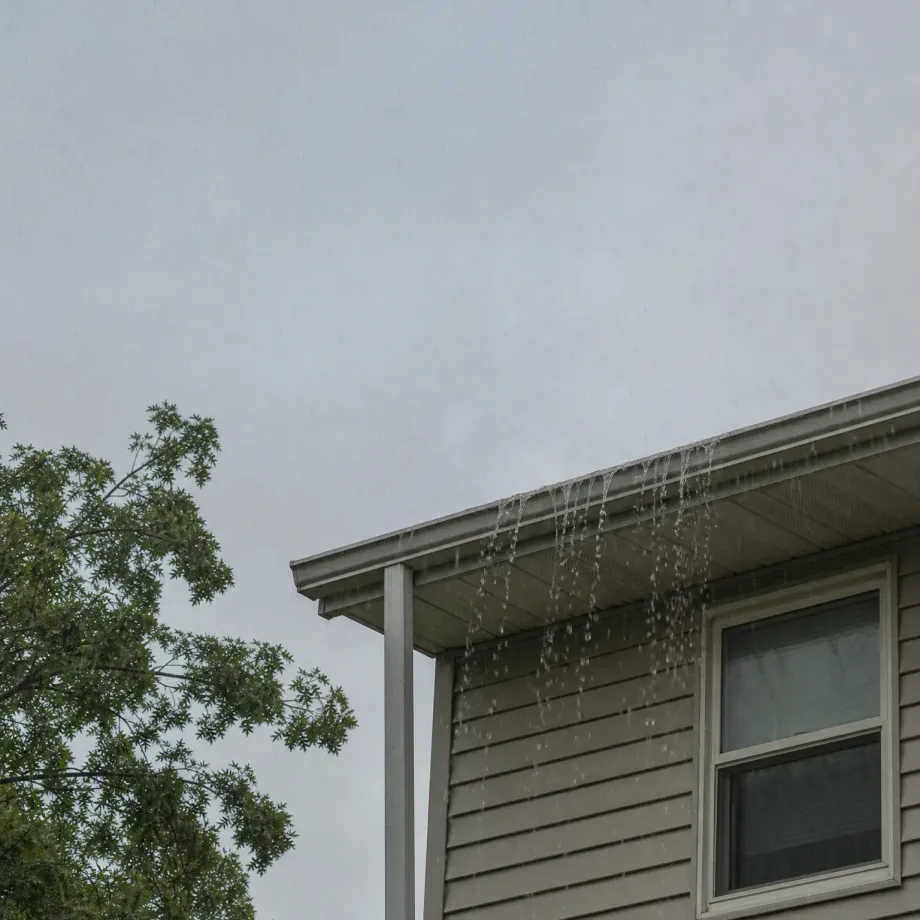 Corner of a house with beige horizontal siding and a white-framed window, with water streaming off the roof eave against an overcast grey sky and a leafy tree at left.
