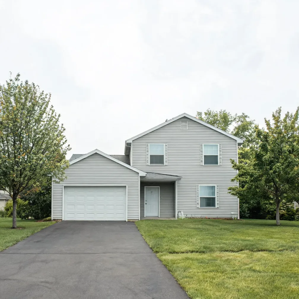 Two-story house with light gray horizontal vinyl siding, a two-car garage, white front door and shuttered windows, fronted by an asphalt driveway, mowed lawn and deciduous trees under an overcast sky.