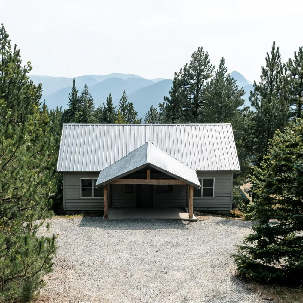 A single-story gray house with a metal gable roof and covered central entry porch, fronted by a gravel drive and flanked by pine trees, with hazy mountain ridges in the background.
