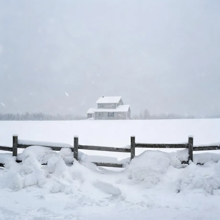 A two-story light-colored house with snow-covered roofs stands beyond a snow-filled field, with a weathered wooden post-and-rail fence half buried in drifts in the foreground under an overcast, hazy sky.