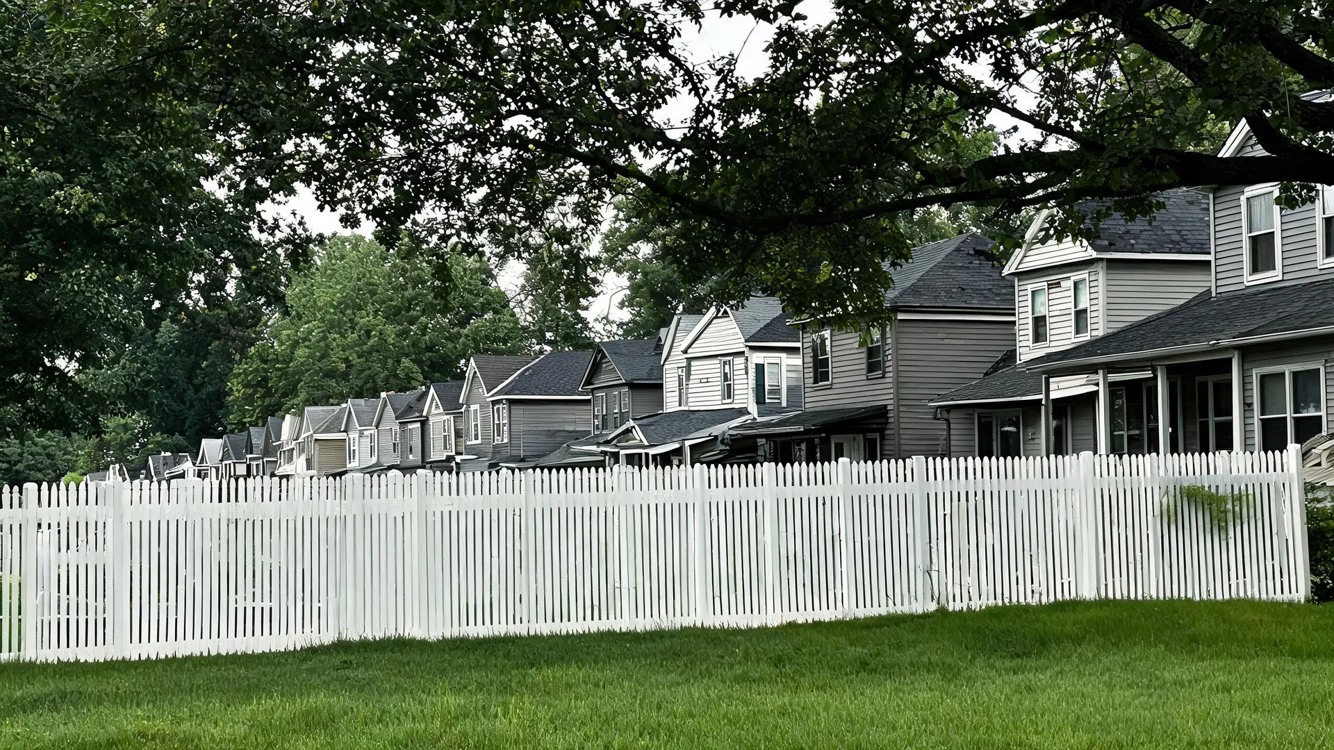 A white picket fence runs along a mowed grass lawn in front of a row of gray clapboard houses with dark shingled roofs, framed by overhanging tree branches under an overcast sky.