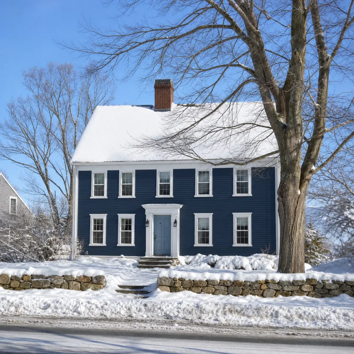 A two-story dark blue clapboard house with white trim, a brick chimney and snow-covered roof, behind a stone retaining wall and snow-covered yard with bare trees.