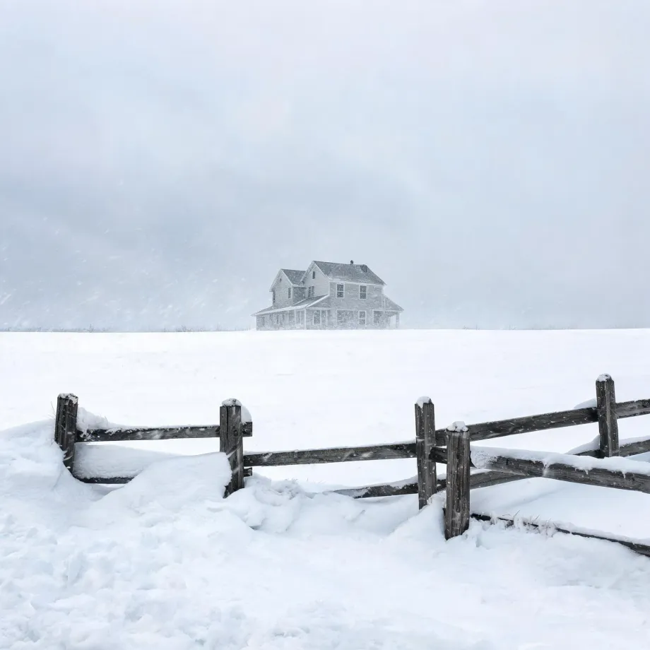 A two-story house with a porch stands alone on a snow-covered field during falling snow, with a weathered wooden split-rail fence partly buried in drifts in the foreground.