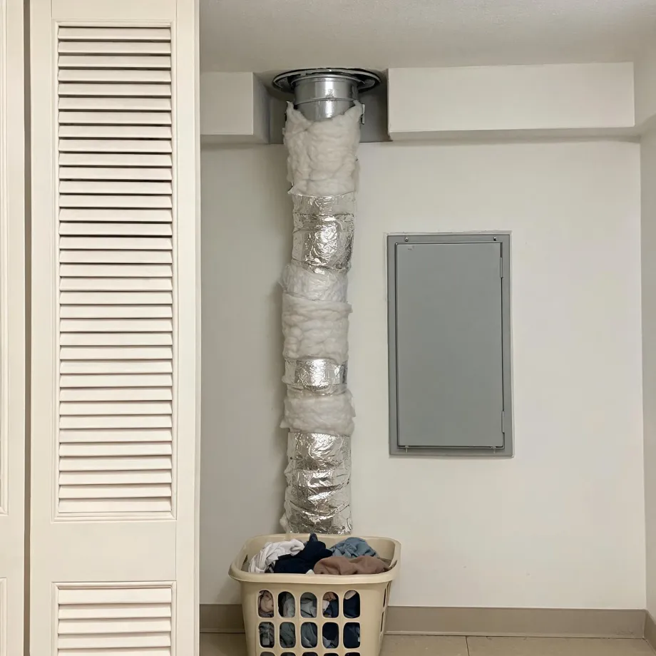 Interior corner of a room where a flexible foil duct runs from a ceiling collar down the wall, partly wrapped with loose white insulation, next to a louvered closet door and a gray wall access panel, with a laundry basket of clothes on the tile floor below.