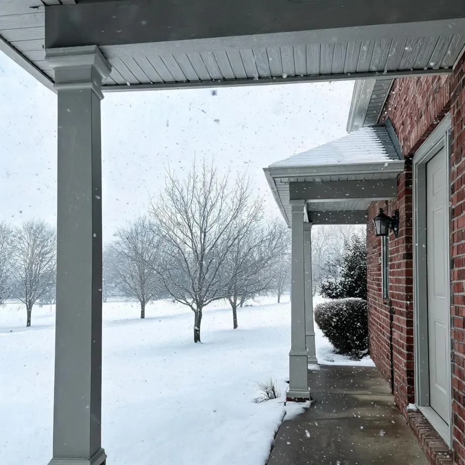 View from a covered porch with white columns of a red brick house during falling snow, looking out over a snow-covered lawn with bare trees and a trimmed shrub beside a lamp and white door.
