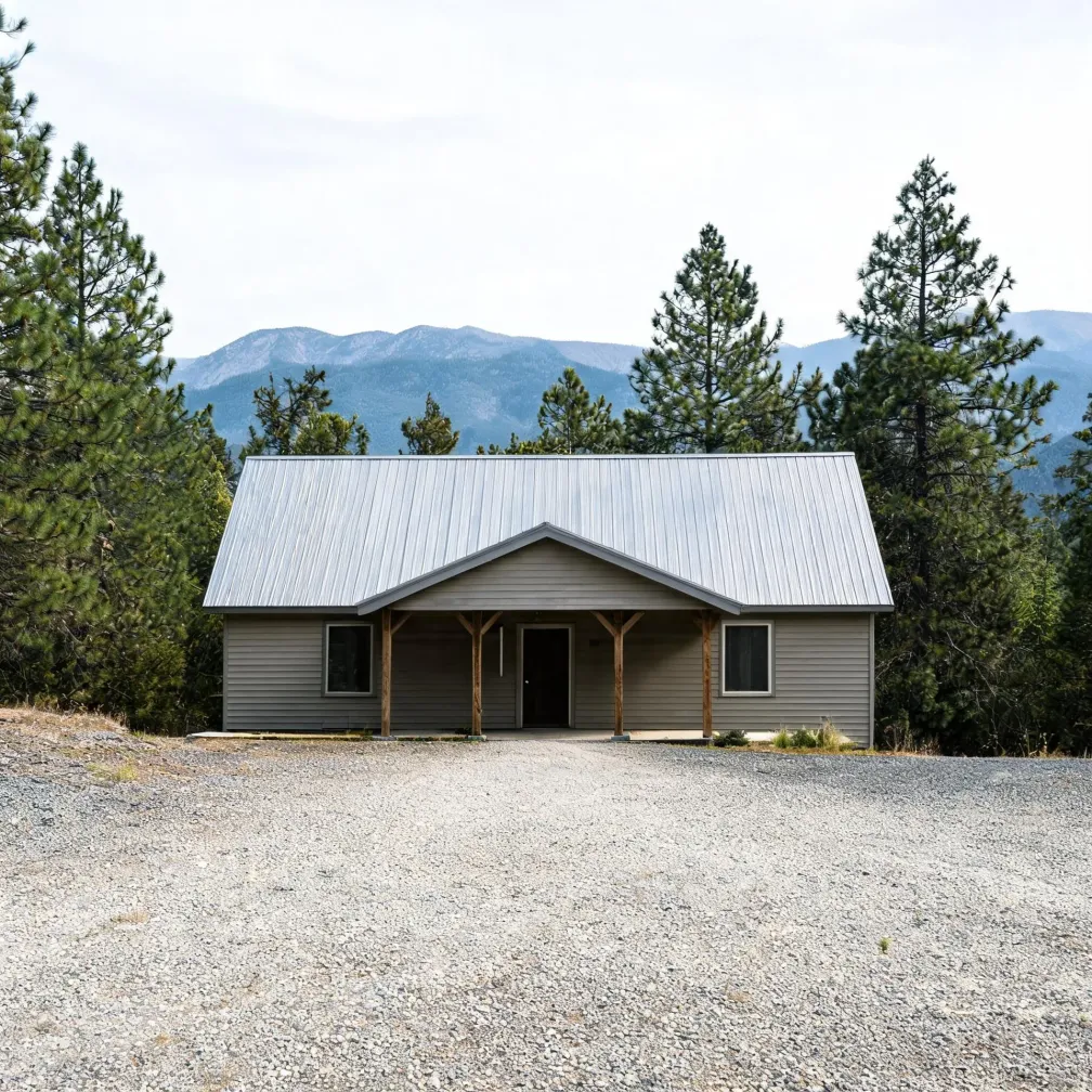 A single-story house with gray horizontal siding and a metal gable roof, with a covered front porch on wooden posts, set behind a wide gravel area with pine trees and distant mountains under an overcast sky.