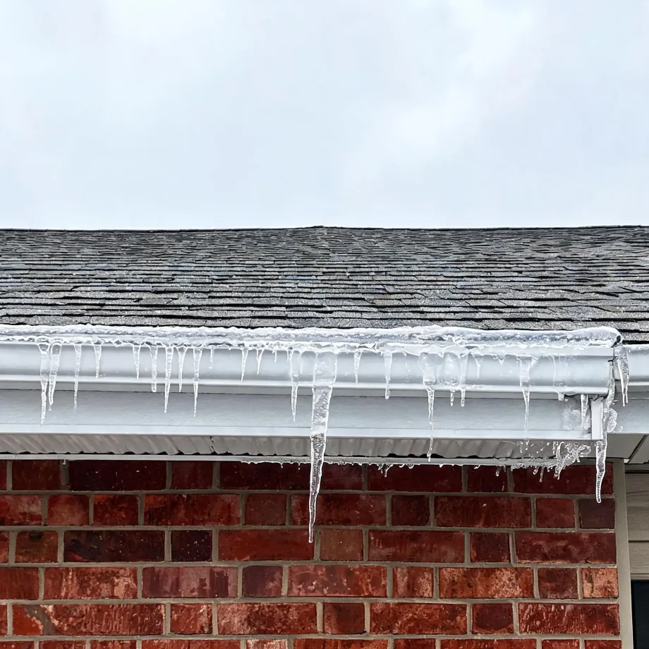 Icicles hanging from a white gutter and soffit below an asphalt-shingle roof on a red brick house, against an overcast sky.
