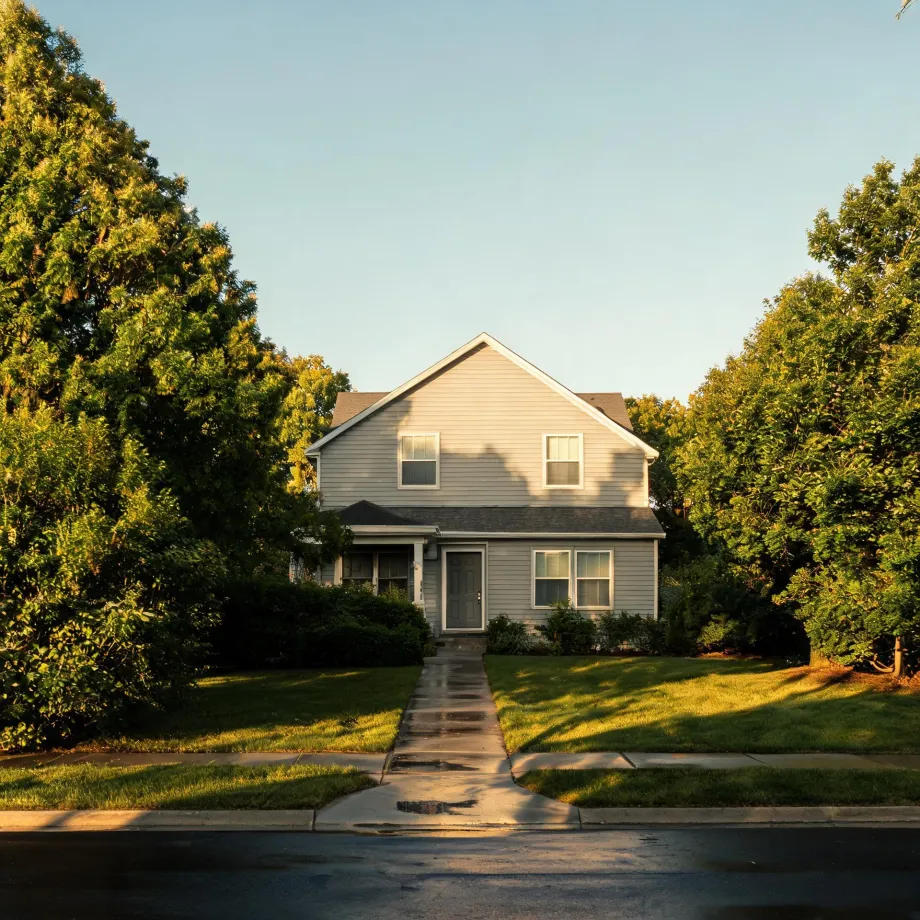 A two-story light gray house with a gabled roof and covered porch, flanked by large green trees, with a concrete walkway crossing a lawn to the street in low sunlight.