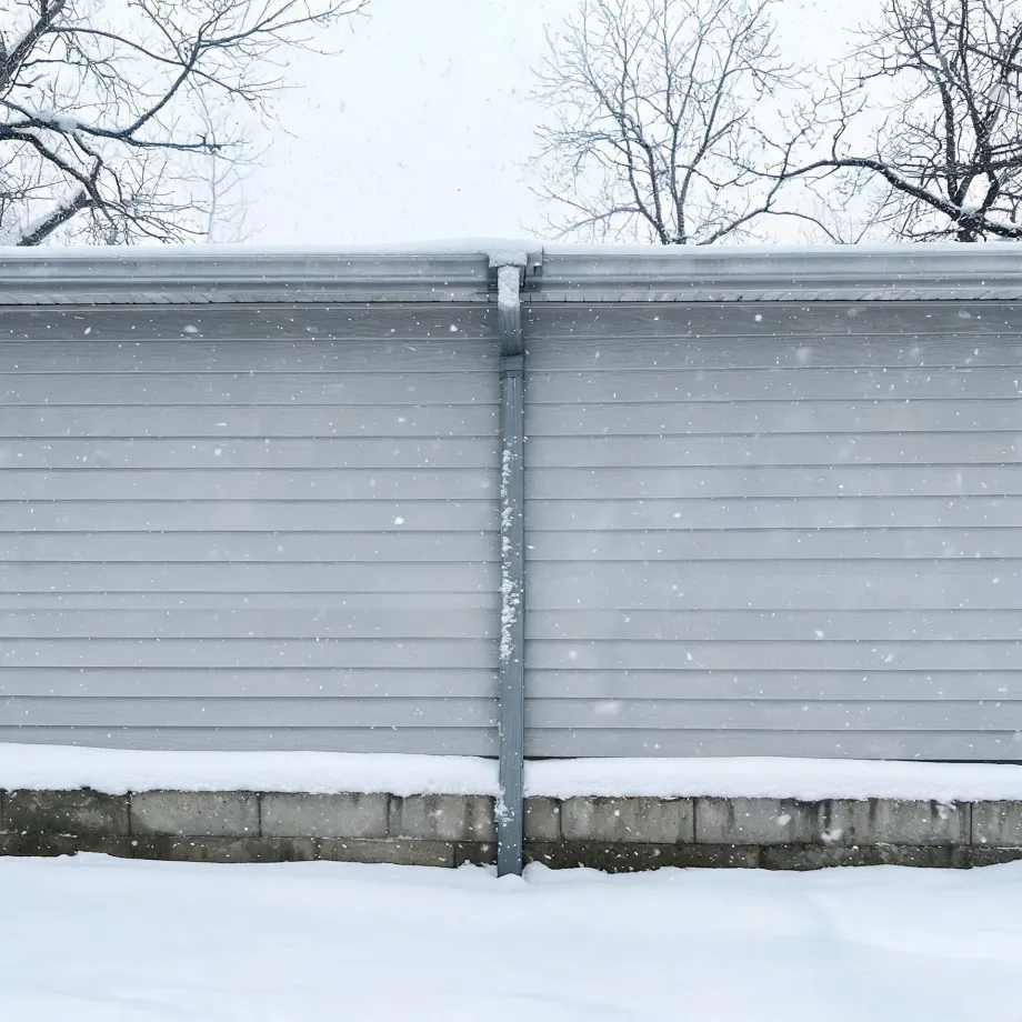 Exterior wall of a building with gray horizontal siding, a metal downspout at center, and a concrete block foundation, with snow on the roof edge, ledge, and ground, and bare trees against an overcast sky during falling snow.