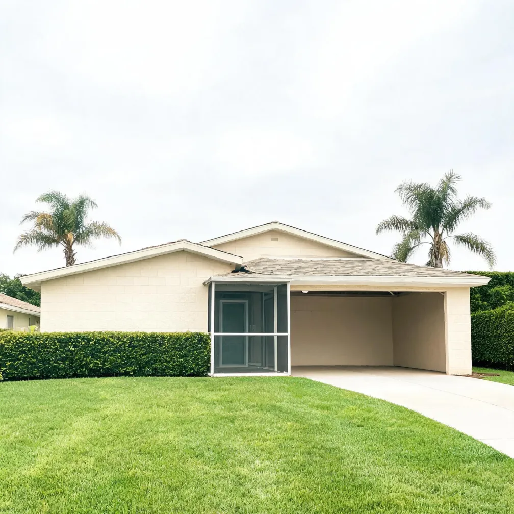 Single-story light-colored stucco house with a shingled roof, an open carport with concrete driveway, a screened entry enclosure, trimmed hedges, palm trees behind the roofline, and a large mown lawn in front under an overcast sky.