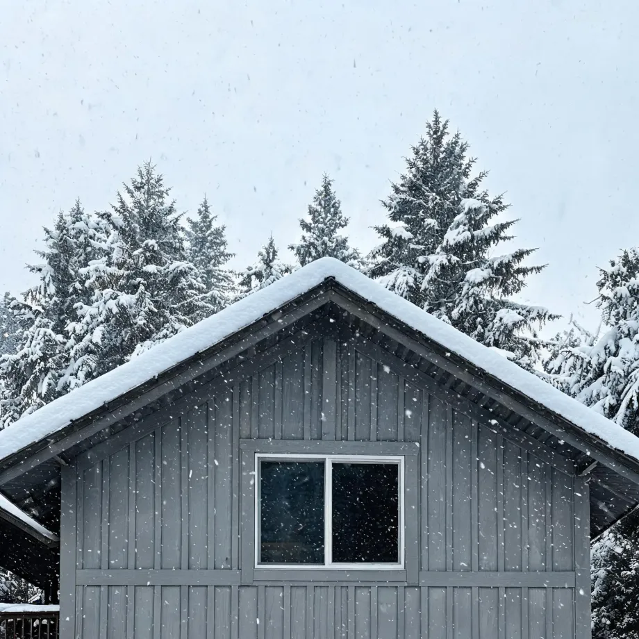 The gray board-and-batten gable end of a house with a snow-covered roof and a single window, framed by snow-laden conifers under a pale sky with falling snowflakes.