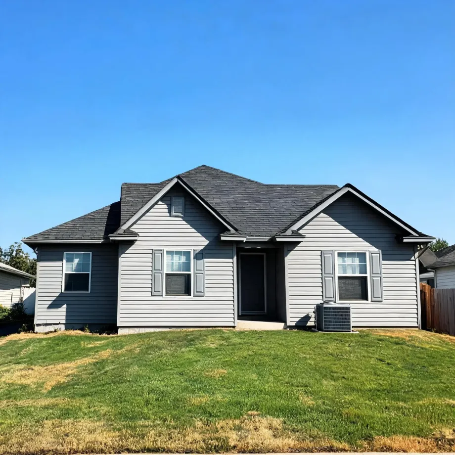 Single-story gray horizontal-siding house with dark asphalt shingle roof, two shuttered windows, a dark front door, and an air conditioning unit, set behind a sloping green lawn under a clear blue sky.