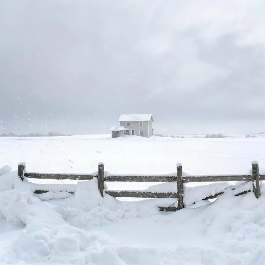 A two-story light-colored house with a gabled roof stands alone on a snow-covered field under an overcast sky, with a weathered wooden post-and-rail fence half-buried in snow drifts in the foreground.