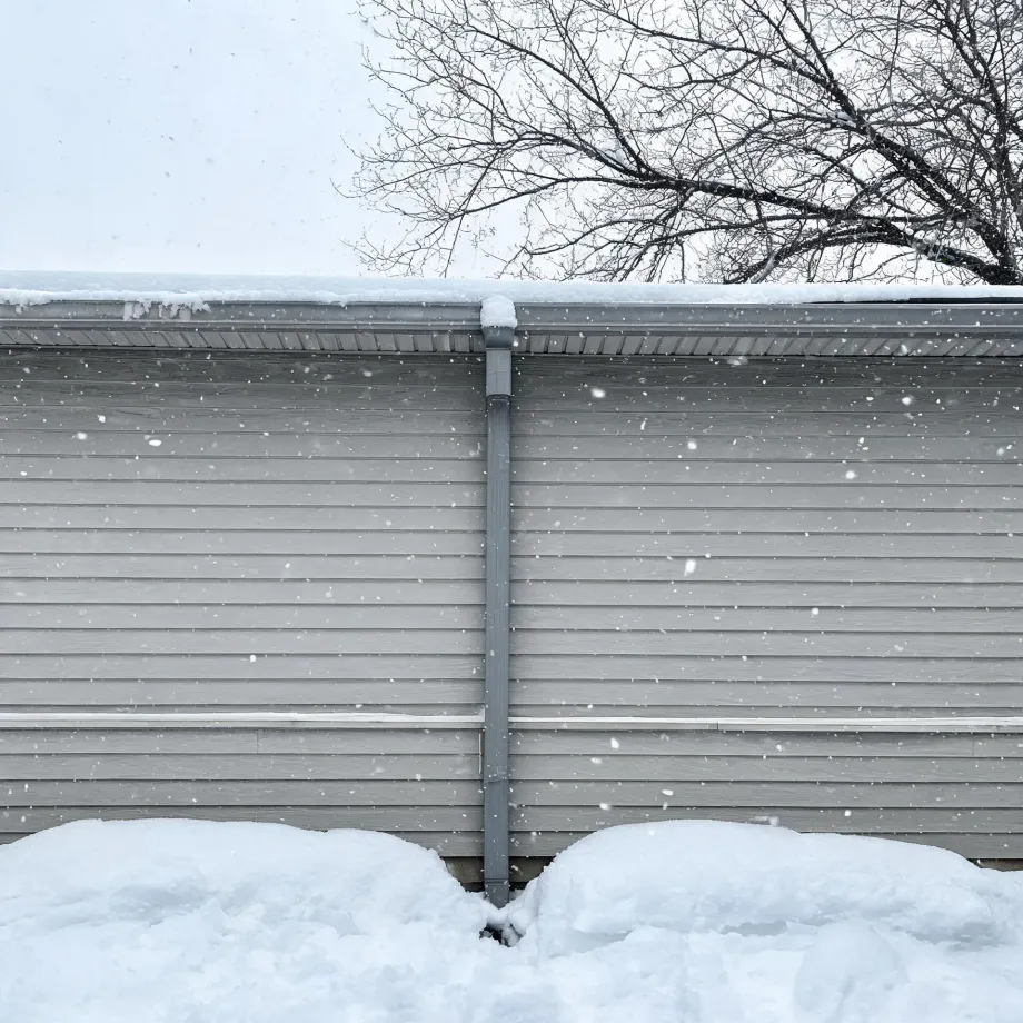 Exterior wall of a building with gray horizontal vinyl siding and a metal downspout, snow piled at its base and on the roof edge, with bare tree branches and falling snowflakes against a pale sky.