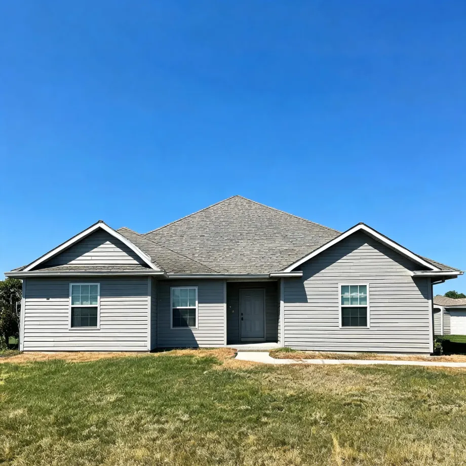A single-story gray horizontal-siding house with two front-facing gables, a shingled hip roof, a dark front door, and three windows, fronted by a patchy lawn and concrete walkway under a clear blue sky.