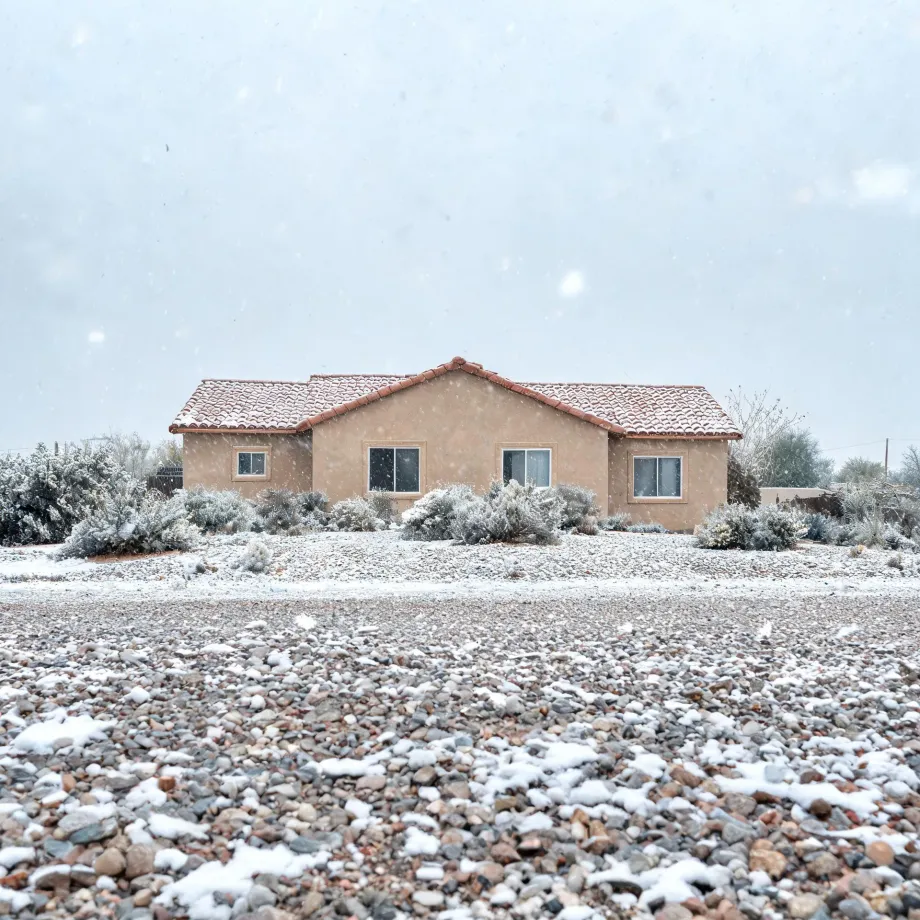 A single-story stucco house with a tile roof behind snow-dusted desert shrubs and a wide gravel foreground during falling snow under an overcast sky.