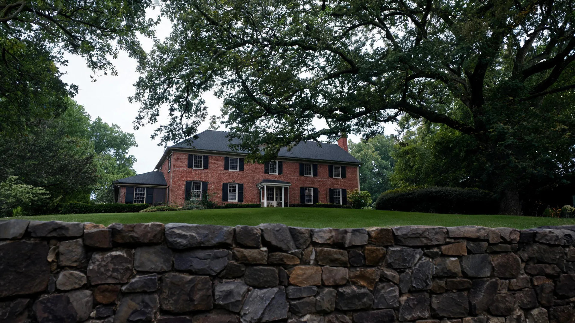 A two-story red brick house with black shutters and a dark roof sits on a mown lawn behind a dry-stacked stone retaining wall, framed by large overhanging tree branches.