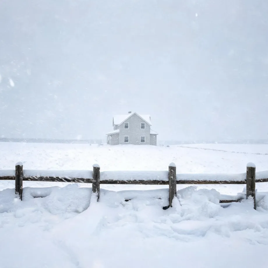 A two-story light-colored house with a gabled roof stands alone in a snow-covered open field during falling snow, with a snow-topped wooden post-and-rail fence crossing the foreground.