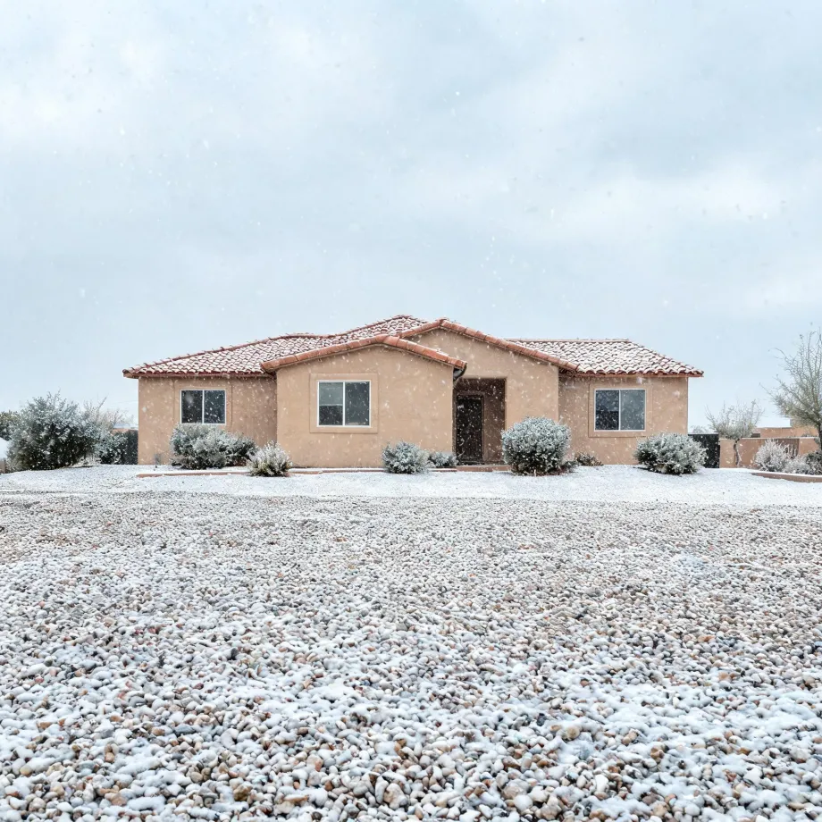 A single-story stucco house with a red tile roof, surrounded by low desert shrubs and a gravel yard lightly covered in snow, under an overcast sky with falling snowflakes.
