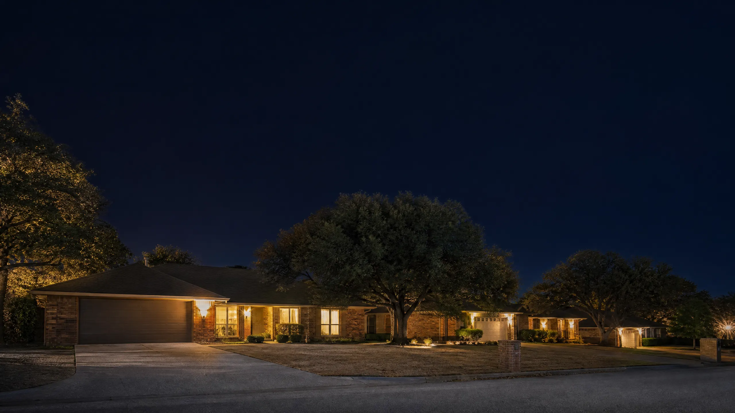 A single-story brick ranch house at night, lit by exterior lights that illuminate the facade, driveway, and several large mature trees in the front yard.