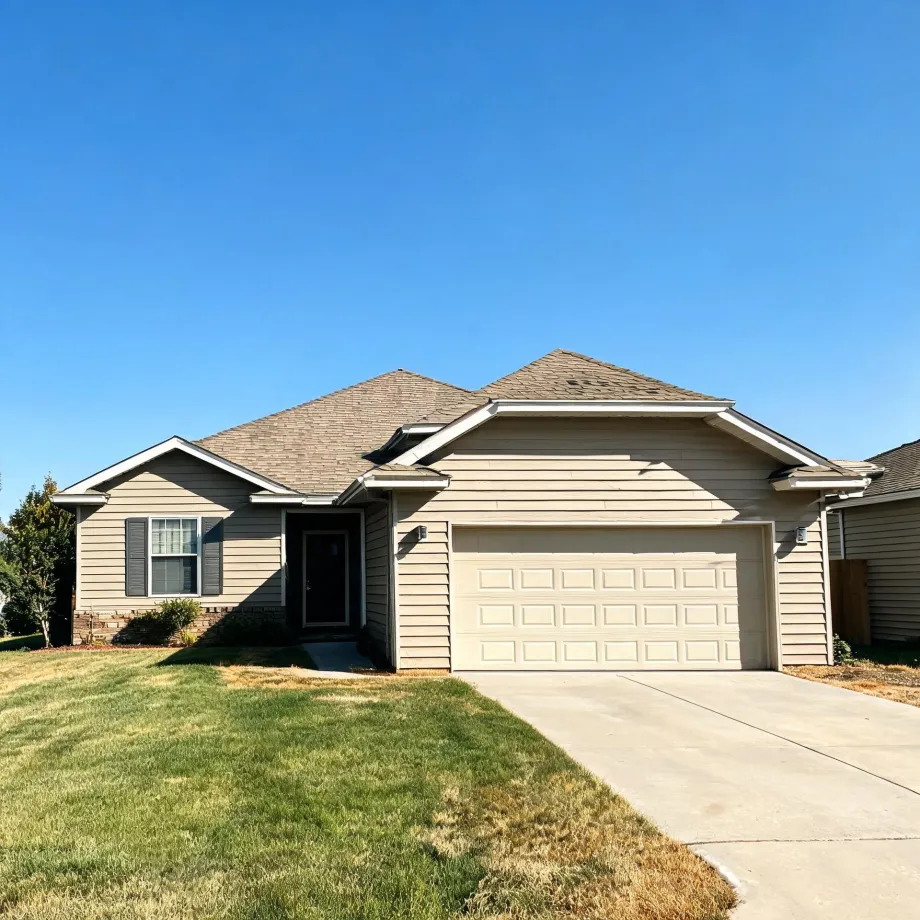 Single-story beige house with lap siding, shingled roof, attached two-car garage with a white door, concrete driveway, and a partly dry front lawn under a clear blue sky.