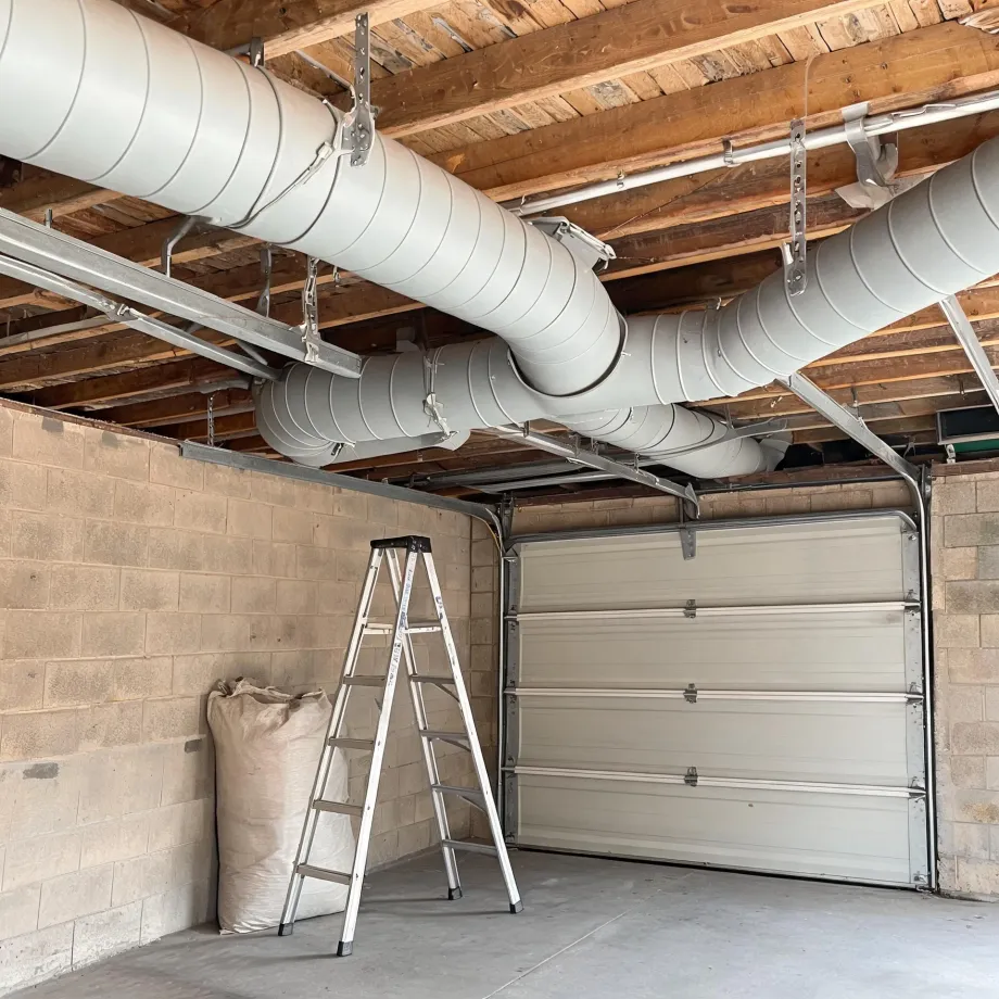 Interior of a garage with concrete block walls, a closed sectional overhead door, exposed wood joists, and large insulated round ducts running along the ceiling, with a step ladder and a filled sack on the floor.