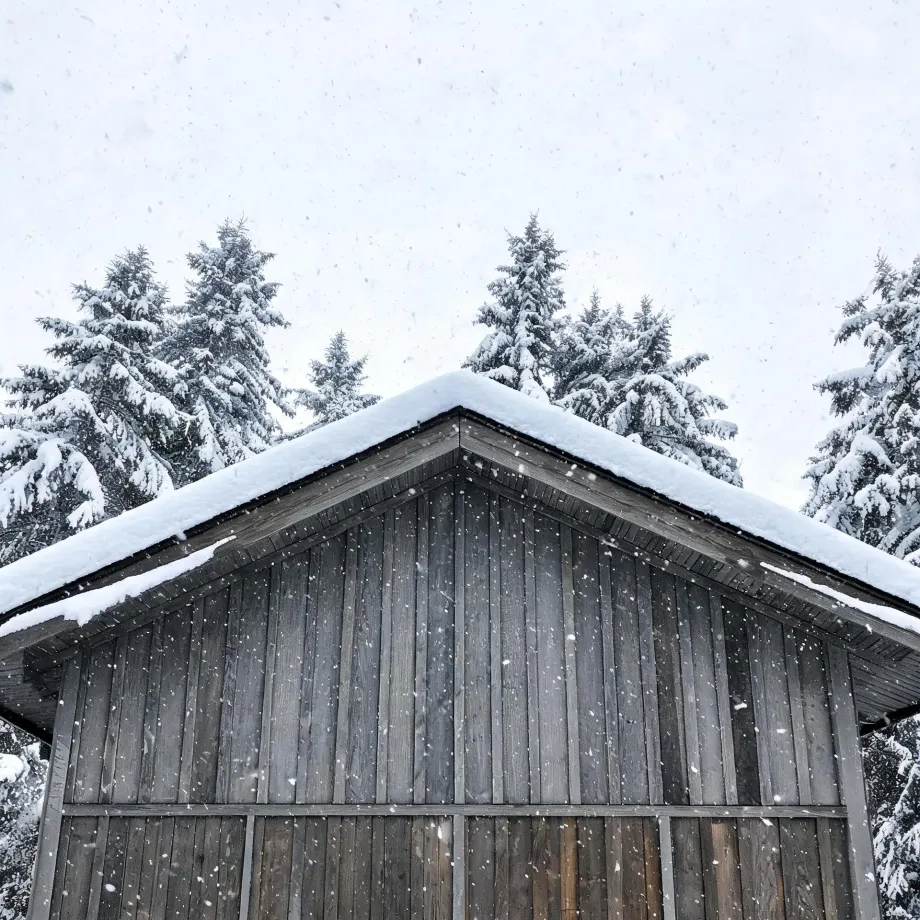 The gable end of a weathered vertical-board wooden building with a snow-covered roof, framed against snow-laden conifers and a pale sky with falling snowflakes.